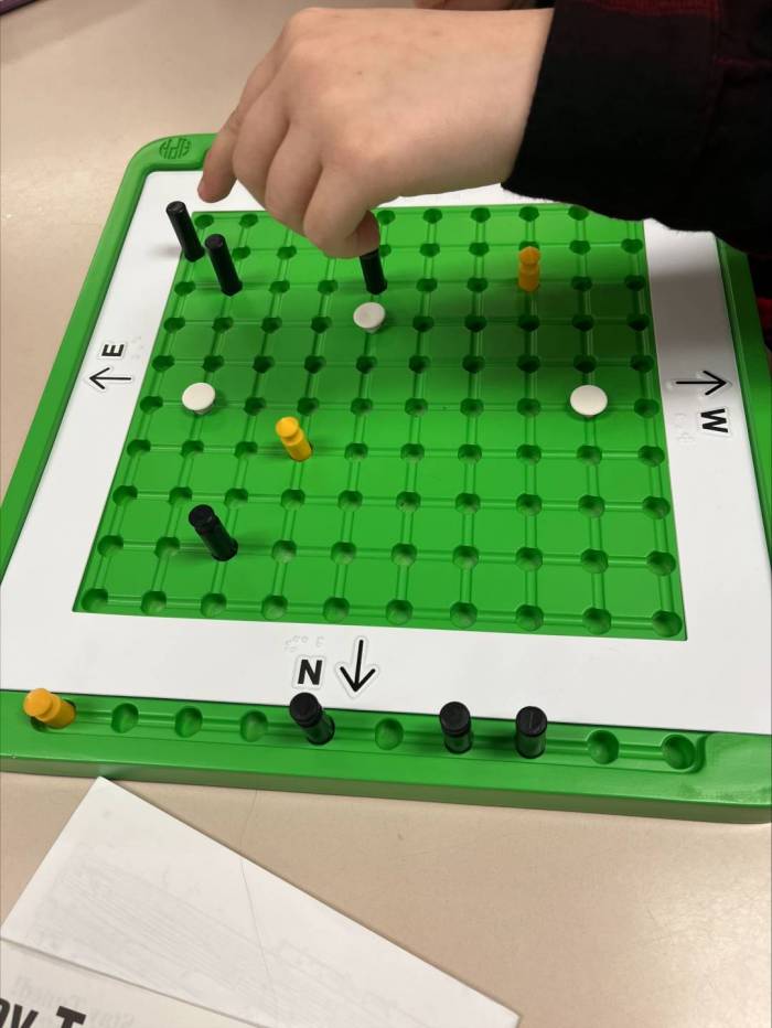 The board game, TREKS, sits on a table. A student's hand can be seen hovering over the board, moving a peg.